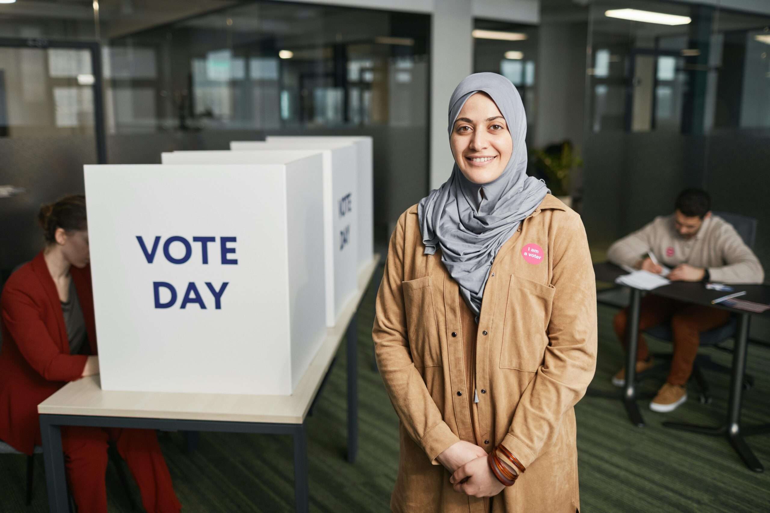 A smiling woman in a hijab stands at a voting booth on election day in a modern indoor setting.